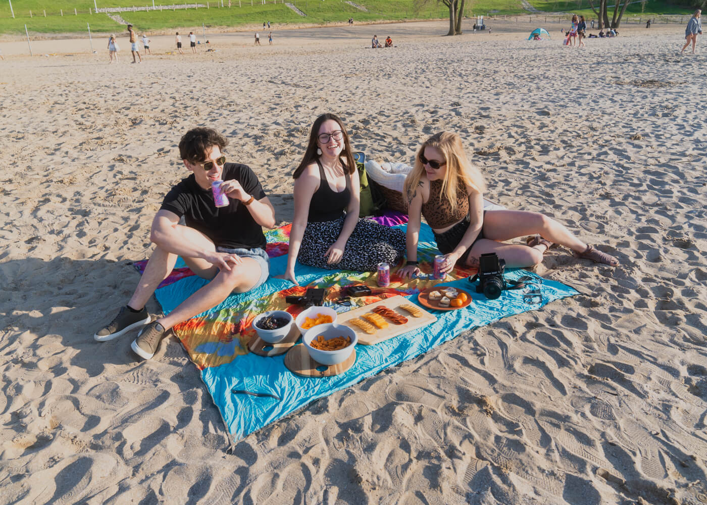 Three people sit on an Islander Blanket at a beach and share a picnic