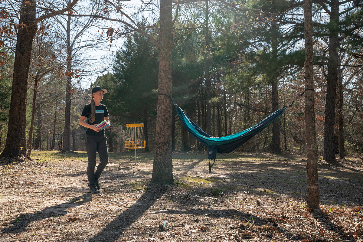 Paige Pierce walks through a park while holding her discs with an ENO hammock and goal in the background.