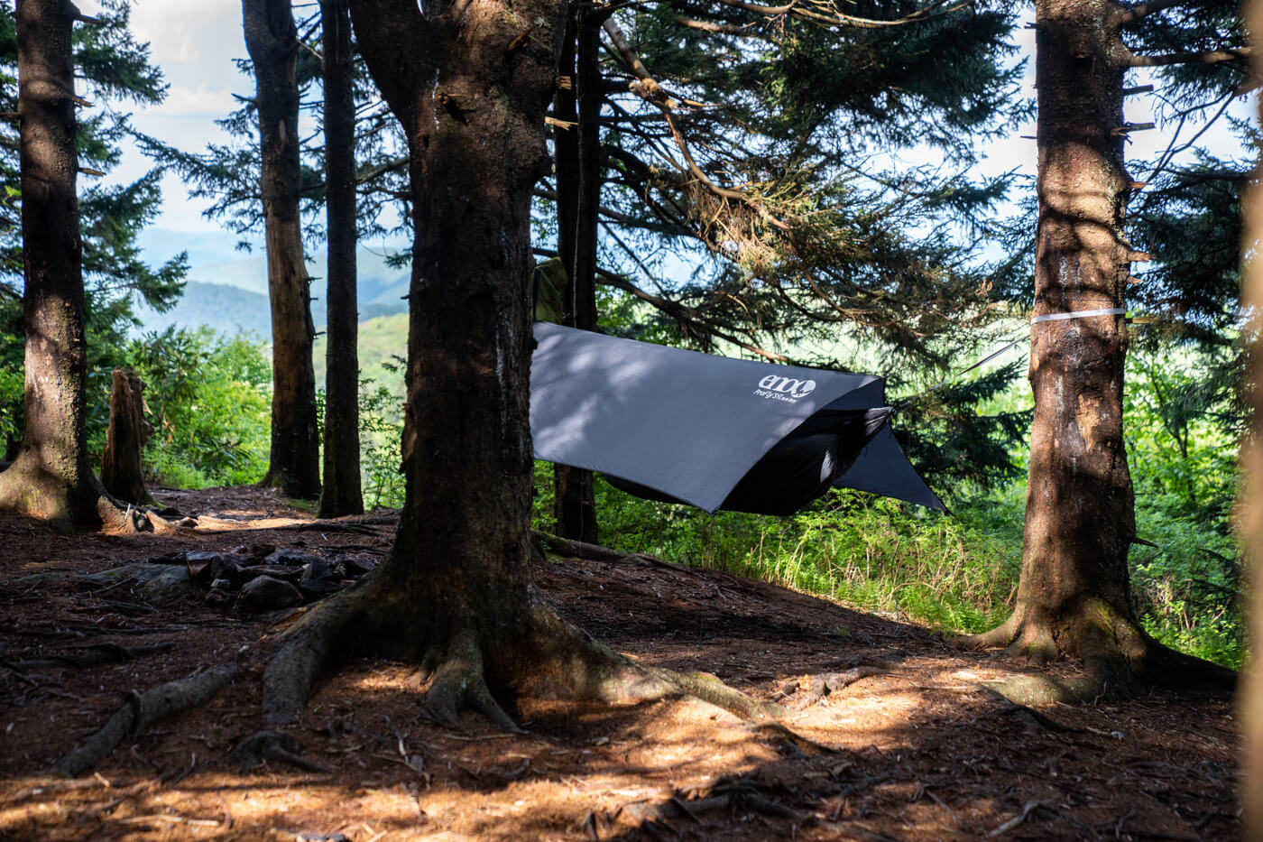 A hammock camping setup is showing in a pine forest with mountain views in the distance. 