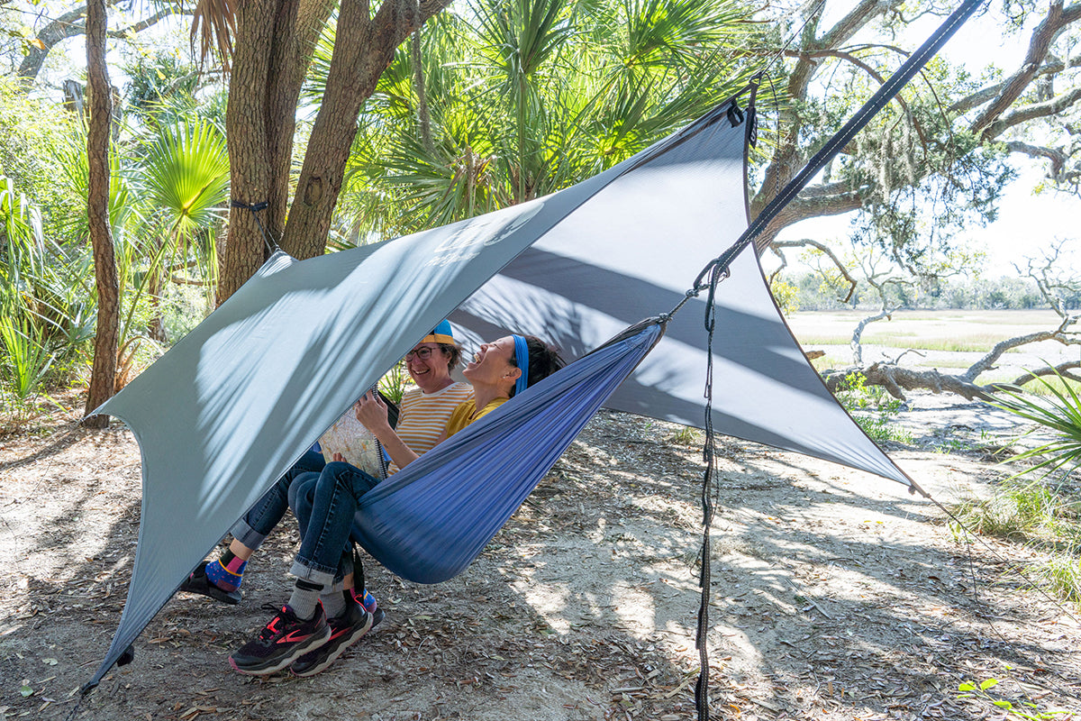 Two women sit together in a hammock under the protection of the ProFly Tarp