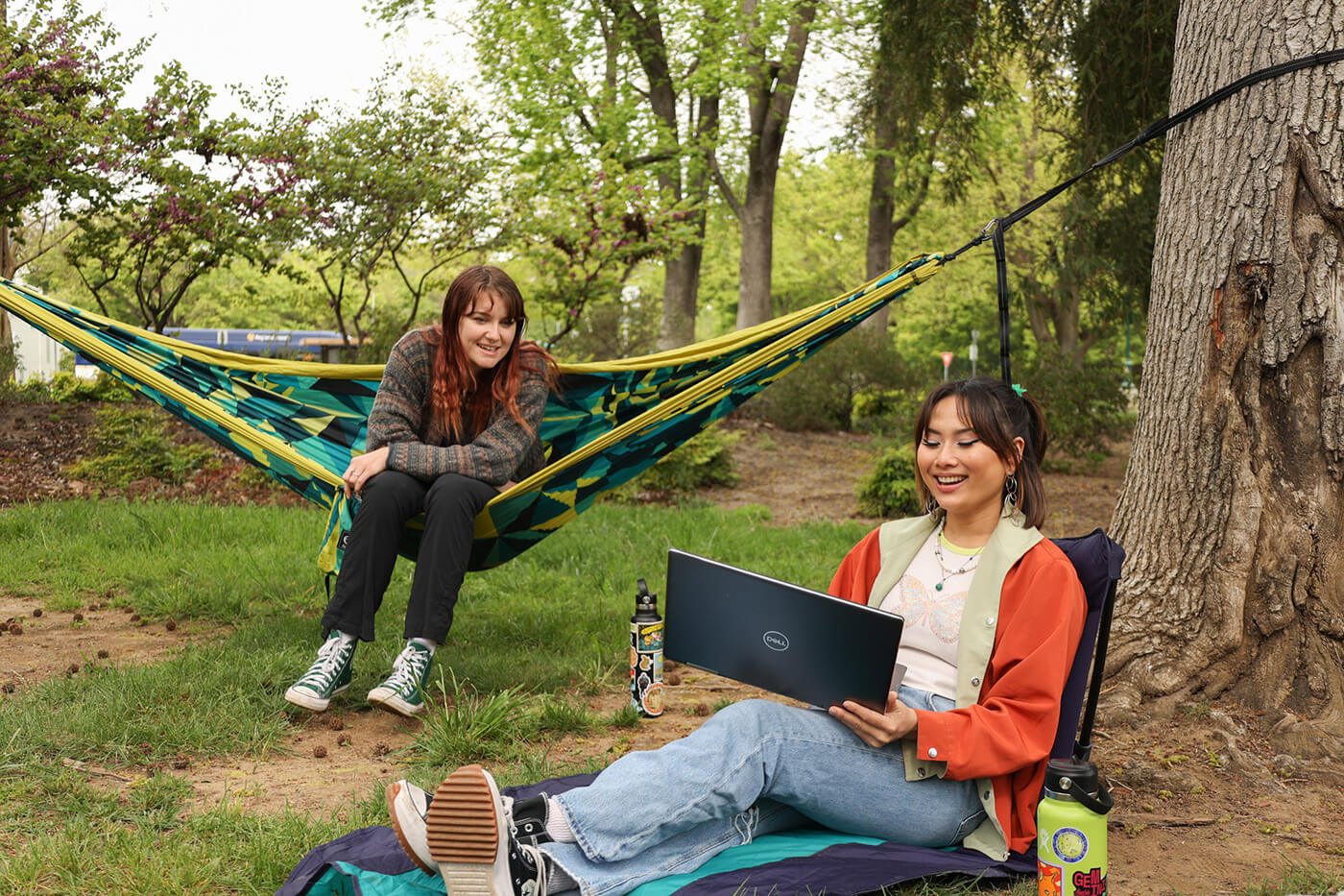 Two college students, one sitting in an ENO DoubleNest Hammock Print and the other in an ENO Lounger GL Chair, look at a computer together on a college gree. 