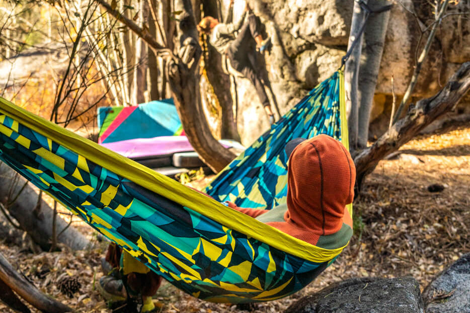A man sits in an ENO Boulder print hammock and watches someone boulder on rocks in the background.