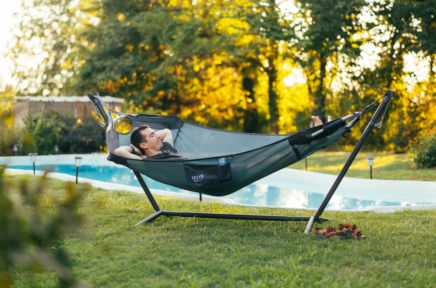 A man relaxes in his DayLoft Hammock and Parkway Hammock Stand while in his backyard, by a pool.