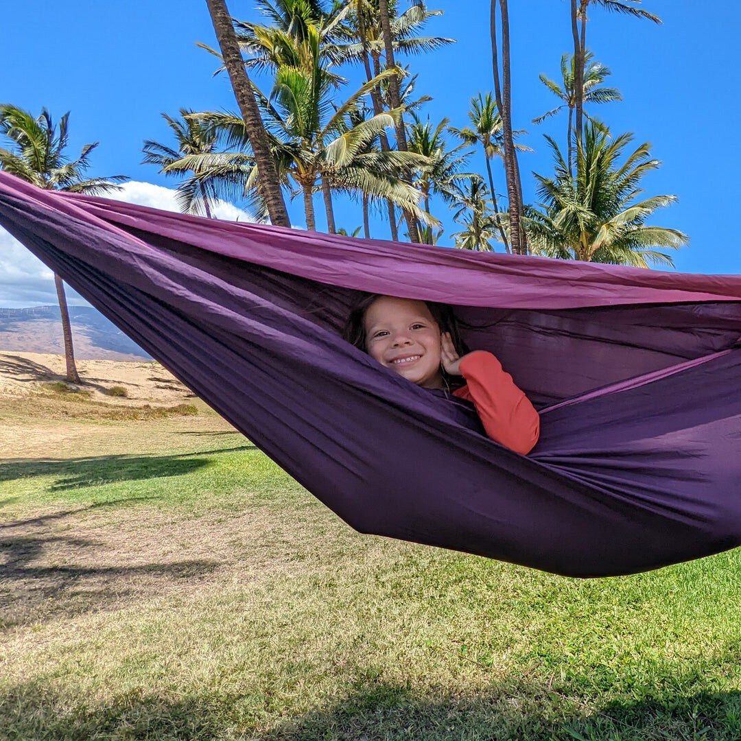 A kid poses for a photo while laying in an ENO hammock in a tropical setting. 