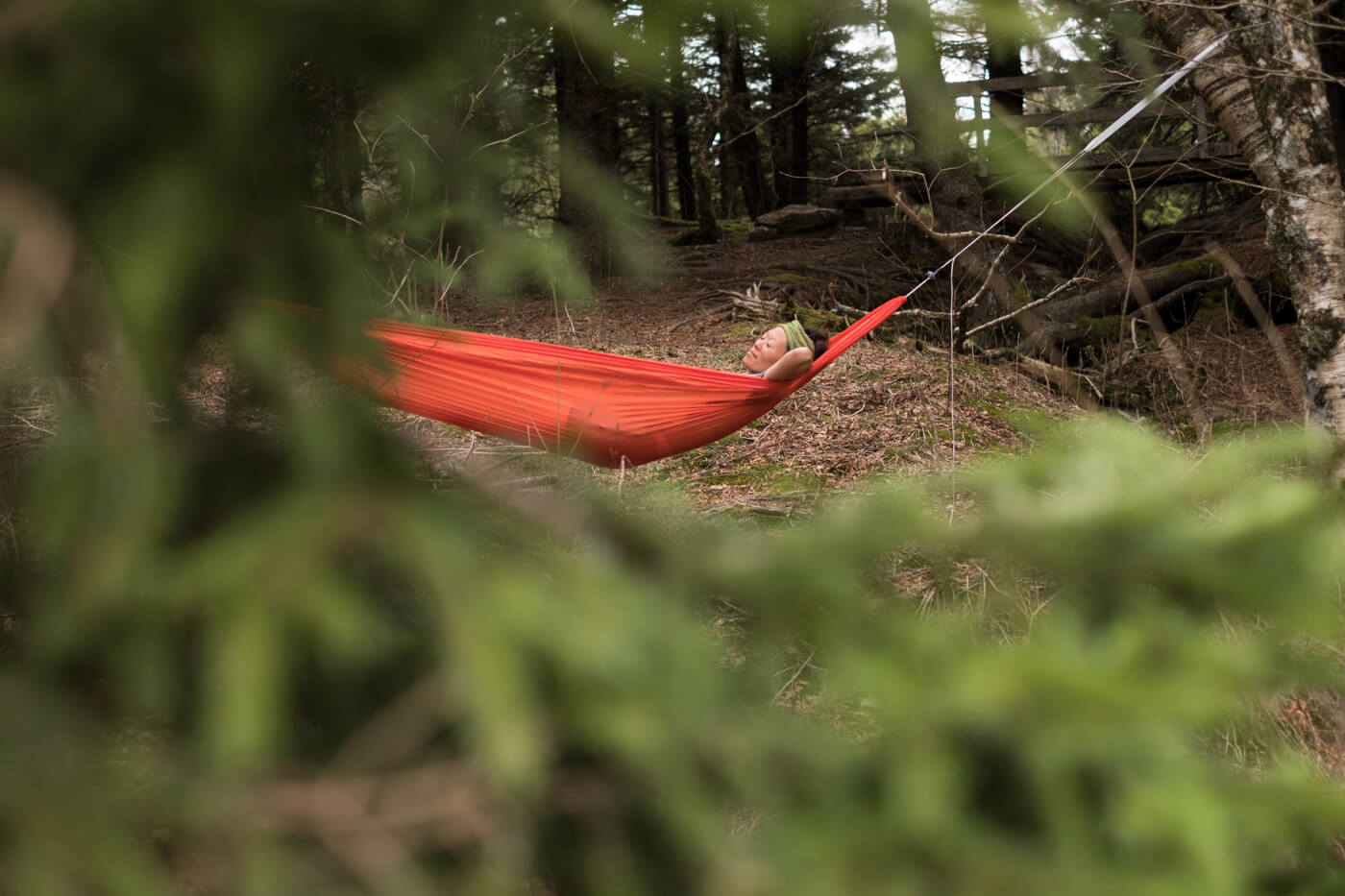 Surrounded by pine trees, a woman lays in a bright orange ENO Sub6 Ultralight Hammock.
