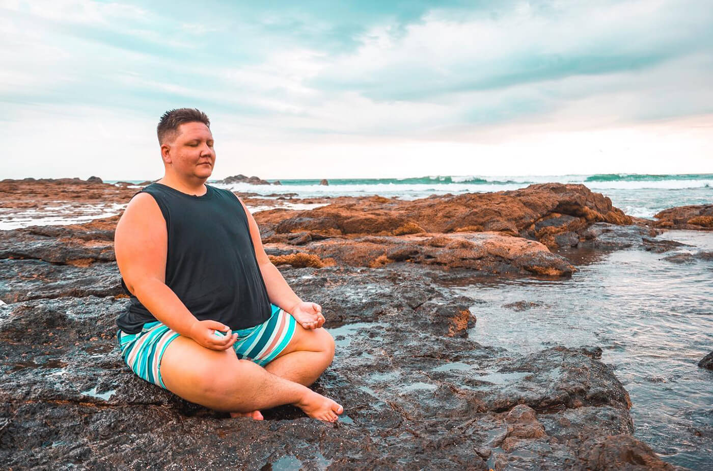 A trans man meditates sitting cross legged on the beach. 