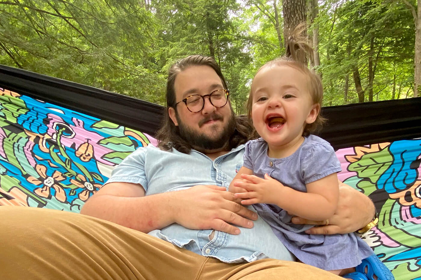 A man hammocks with his daughter in a colorful ENO DoubleNest Print named Nature Talk. 