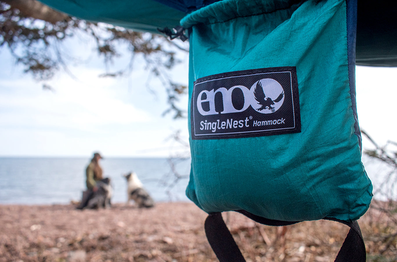 An ENO SingleNest Hammock hangs between two trees while a woman pets her two dogs. 