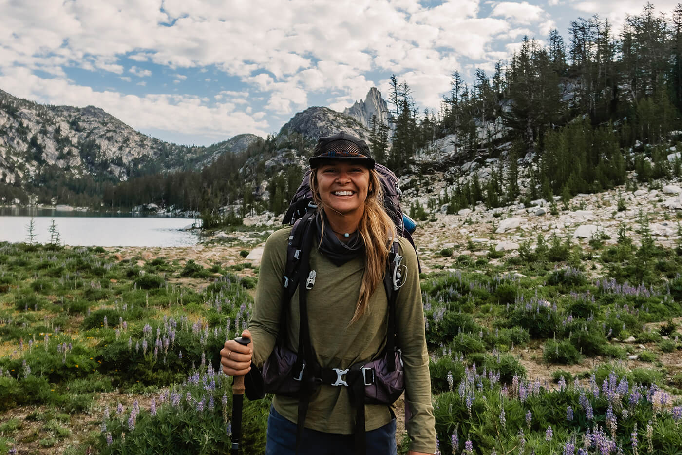 a woman poses for a photo on a trail while backpacking through the mountains.