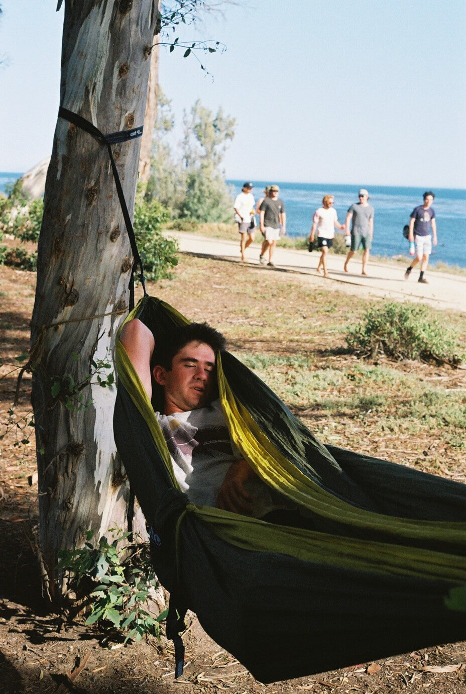 A man lays in an ENO hammock and rests his eyes, with a beach and a boardwalk in the distance. 
