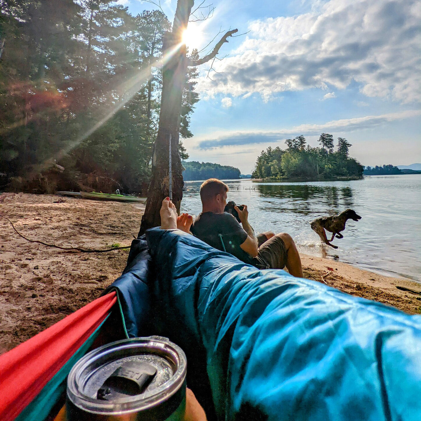 A woman lays in her hammock with a thermos next to her partner holding a camera and a dog running on the beach.