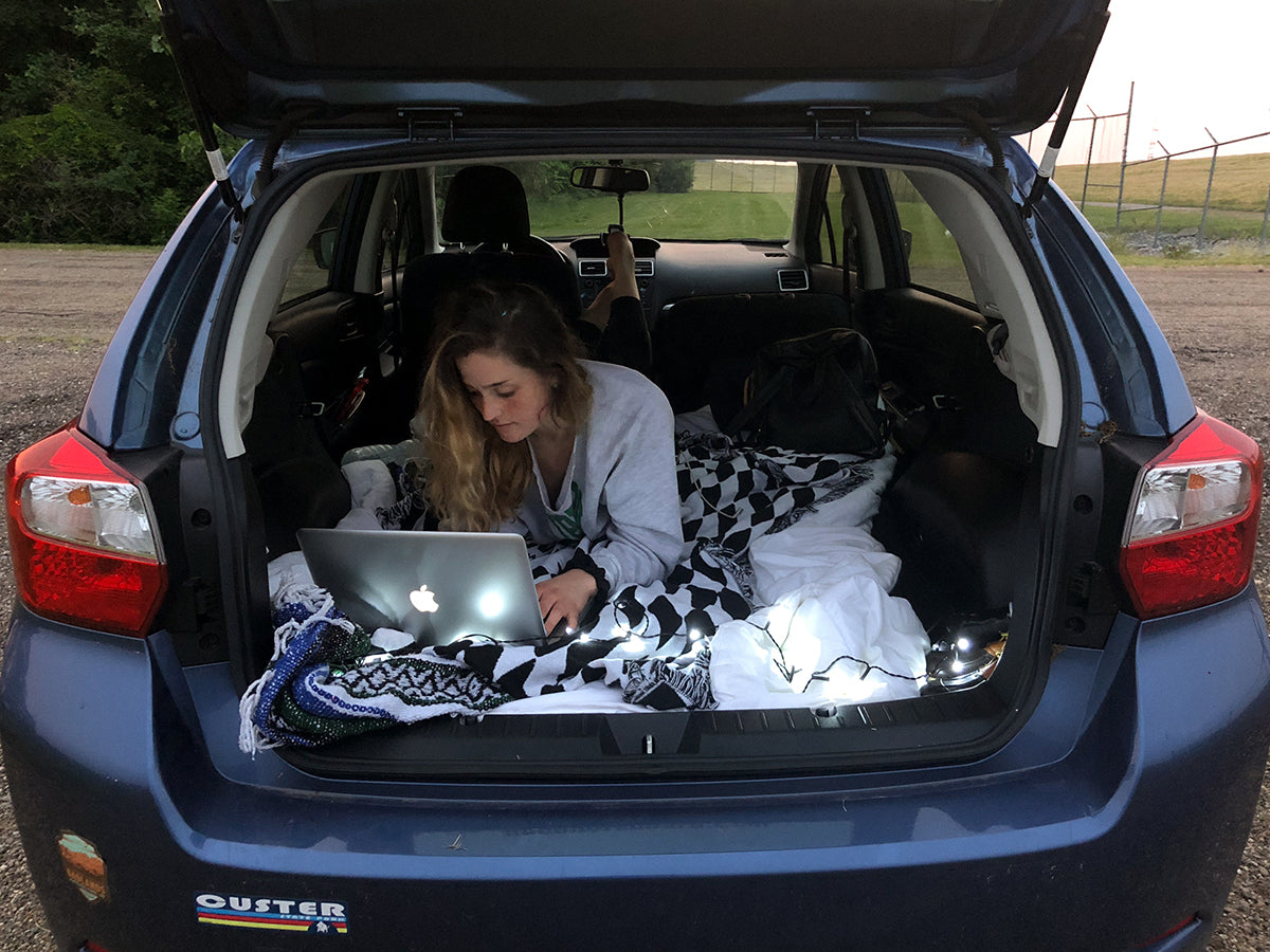 A girl sits in the back of a Subaru while on her laptop.