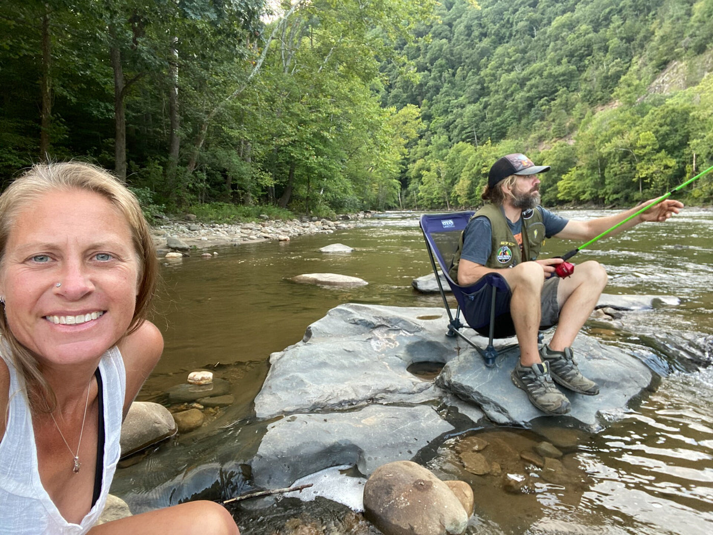 Two people sit in chairs in the creek together and fish, while posing for a selfie. 