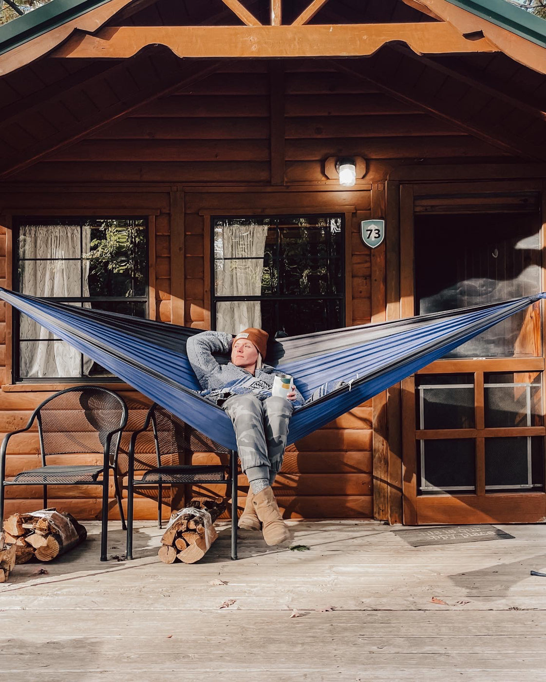 A woman sits in hammock put up between two posts in front of a cabin. 