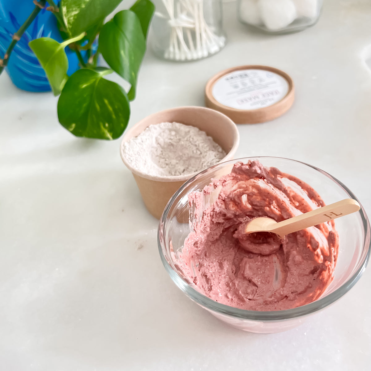 A powedered face mask is mixed into a bright pink paste in a small clear bowl with a small wood spoon.