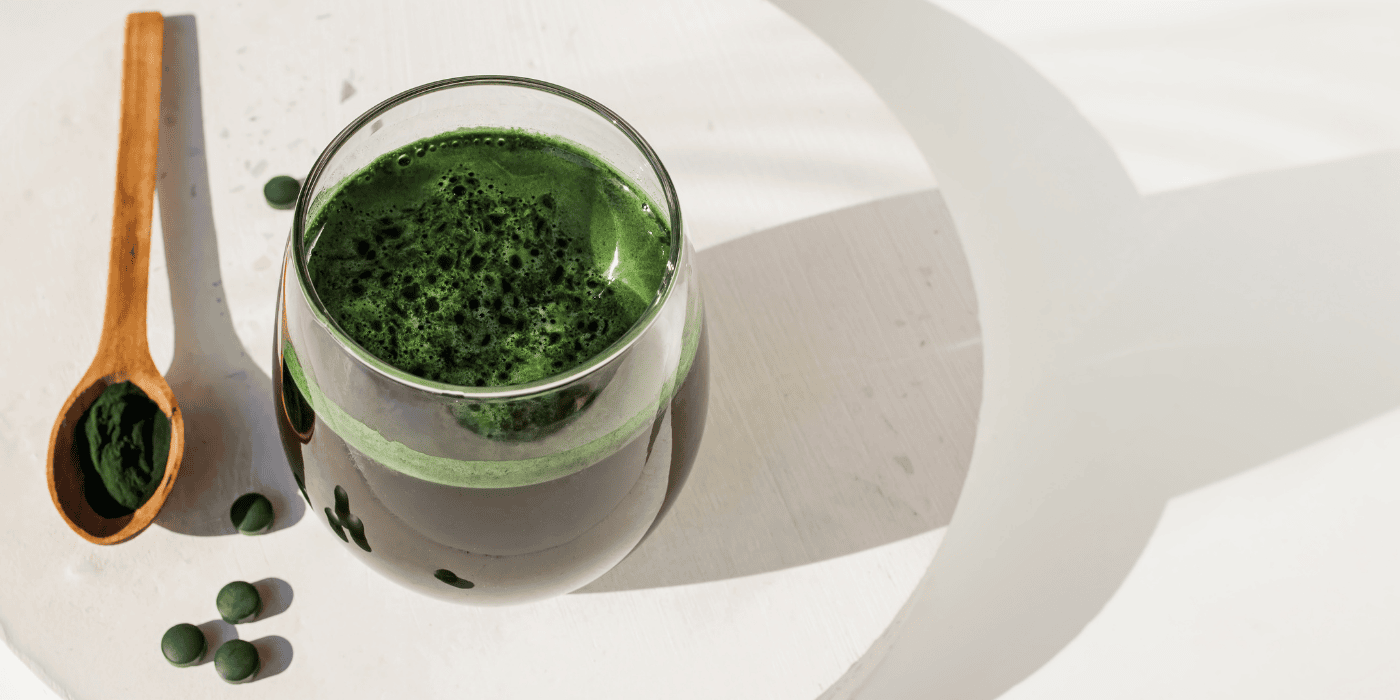 Chlorella-Saft in einem Glas, daneben Chlorella-Pulver in einem Holzlöffel