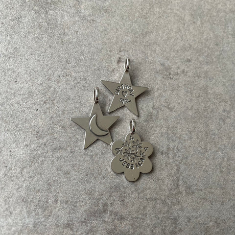 3 silver LifeStory charms set against a grey background. From top to bottom: star shape with the lettering “BIRTHDAY GIRL” and a heart stamp in the centre. Middle: silver star shaped charm with a fused-on moon shape in the centre. Bottom: flower shaped charm with stamped flower pattern and the name “JESSICA” at the bottom.