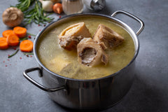 A silver stock pot with handles on a gray counter top, full of broth with 4 beef shin bones visible, and carrots, garlic, and onions in the background