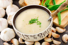 A bowl full of off-white colored soup with chives in the center, surrounded by several heads and individual cloves of garlic