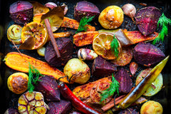 A collection of colorful, roughly sliced, browned vegetables, including beets, garlic, carrots, lemons, and sweet potatoes, garnished with fennel fronds.
