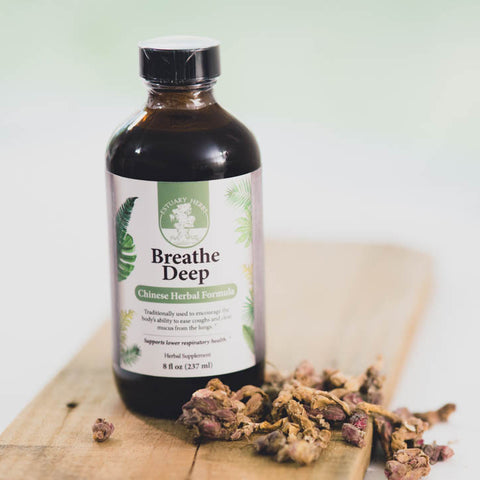 An amber glass bottle with a product label, atop a cedar plank, with a small pile of an herbal ingredient in front, on a blurry white and green background.