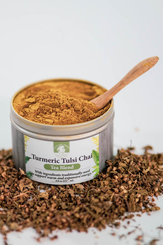 A cylindrical tin full of an orange-brown powder, with a small bamboo spoon resting inside, surrounded by tiny, brown, dried tulsi leaves, on a grayish background.