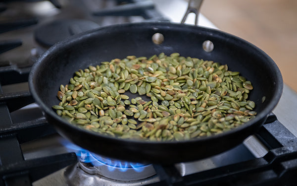 In a dry skillet, lightly toast the pumpkin seeds over medium low heat, stirring regularly, until they smell toasty and just start to change color and pop, approximately 7 minutes. 