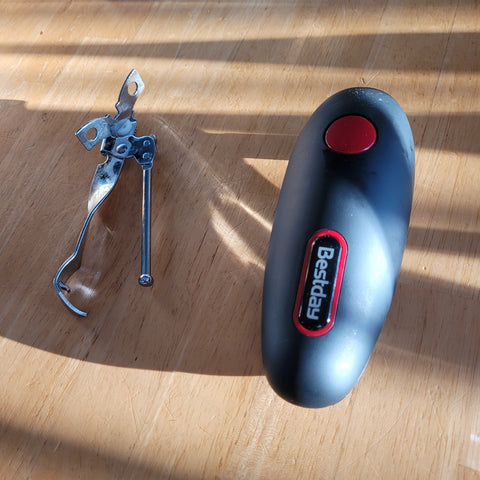 A metal, very traditional can opener sitting on a table next to a black electric can opener with a red button.