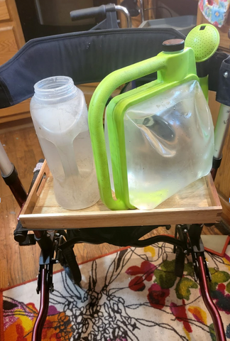 A green watering can with a collapsible clear plastic "belly" filled with water is sitting on a walker.