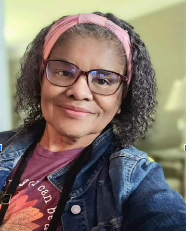 Karen, a black woman with grey hair wearing glasses and a pink headband, is smiling at the camera. She's wearing a maroon tshirt and jean jacket