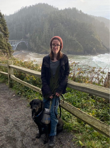 Mary is outside on a mountain trail leaning against a fence. She is wearing glasses and has shoulder-length brown hair. She is wearing a red beanie. Beside her is a black lab looking dog, Amara. Behind Mary is a river and a bridge.