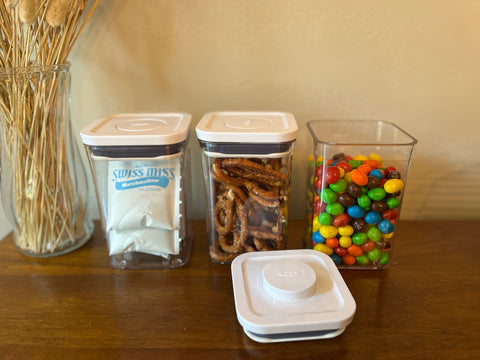 Three clear containers on a wood table, one filled with hot chocolate, a second with pretzels and a third with colorful m&ms.