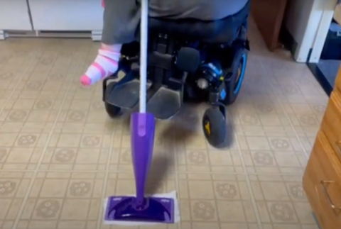 Purple Swiffer Wet Jet being used from a power chair on a linoleum kitchen floor.