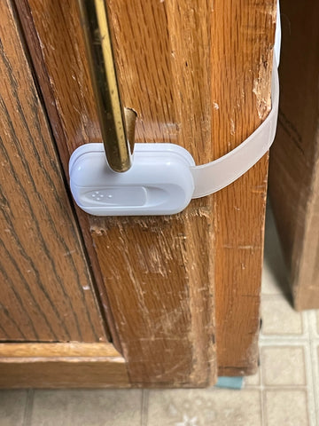 A white baby lock is attached to the door of a cabinet and holding it shut with a strap fixed to the side of thee cabinet.