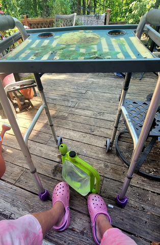 Now the flexible watering can is sitting at the feet of a person wearing pink crocs. A walker is on an outdoor deck in front of the person, where the watering can will go..