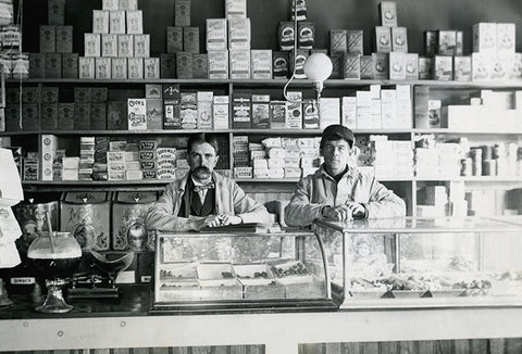 An old, black-and-white photograph of two men standing behind the counter in a grocery store during wartime.