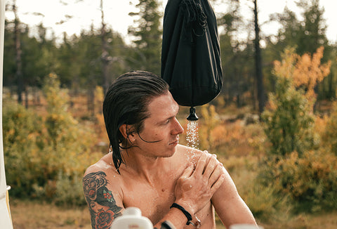 A tattooed man taking an outdoor shower with a rain poncho in the wilderness.