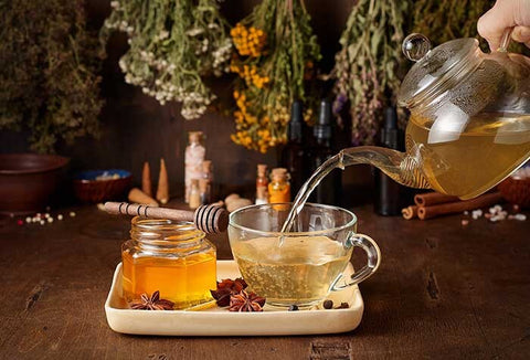 Someone pouring a teapot of tea into a teacup. The teacup is placed on a white tray, next to golden honey.