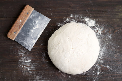 Raw, round bread dough in flour next to a bread cutter on a dark wood table.