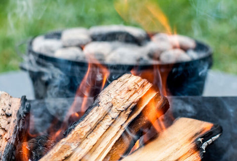 Firewood on fire with a dutch oven covered with coals in the background.