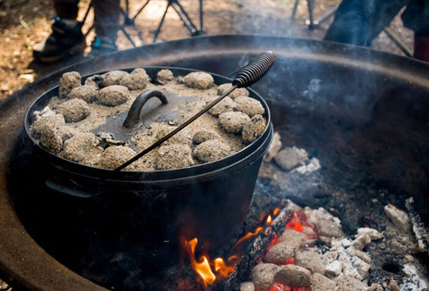 A dutch oven covered with coals sitting in a campfire outdoors.