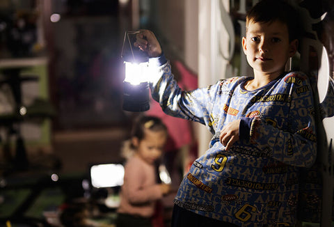 A young boy holding an electric lantern inside a dark home with his mom and little sister behind him.