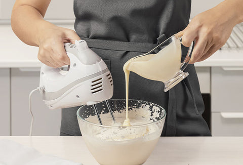 Person pouring cream into a glass bowl while holding a handheld electric mixer.