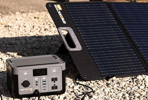 Compact generator next to a solar panel angled toward the sun, both sitting on gravel.