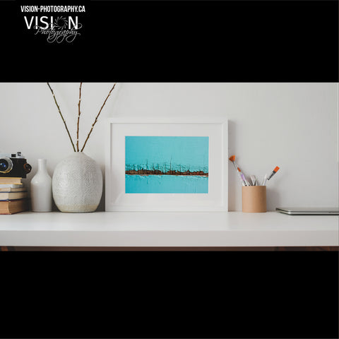 A desktop that has a small framed abstract photo that is robin egg blue and rusty wood. To the left of it is a vase and to the right of it is a jar with paintbrushes. The art is created by Laura Cook of Vision Photography. 