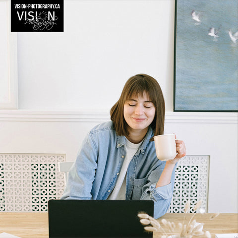 a relaxed lady in her home office, sitting behind a small laptop with a coffee in her hand. She is smiling and on the wall is a picture of 'Freedom Flight' an abstract of white bird on a blue lake. The blue in the photo matches the ladies light blue denim shirt. The art is created by Laura Cook of Vision Photography