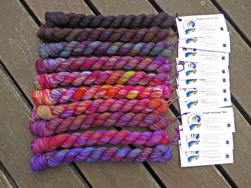 Ten variegated mini skeins of yarn in shades of grey, mauve and purple with white labels are arranged from darkest grey at top to lightest grey then through mauve  to purple at bottom, lying on a brown wooden background.