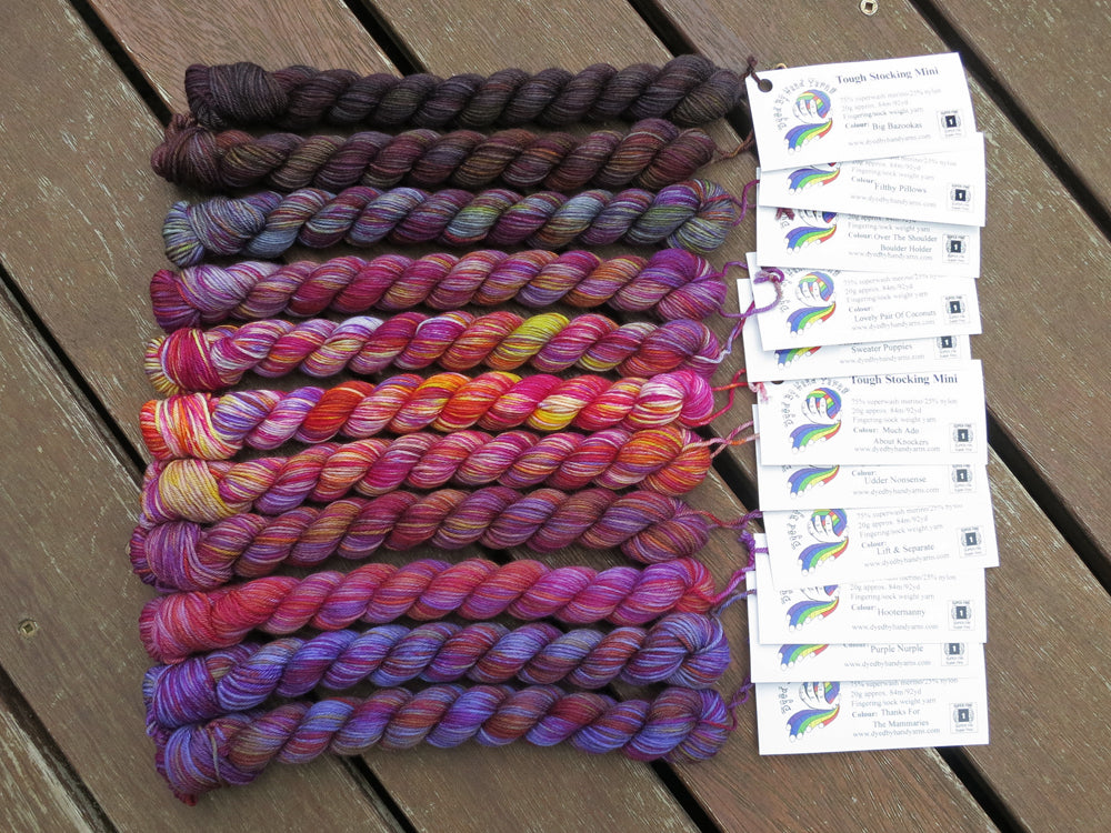 Eleven variegated mini skeins of yarn in shades of grey, mauve and purple with white labels are arranged from darkest grey at top to lightest grey then through mauve  to purple at bottom, lying on a brown wooden background.