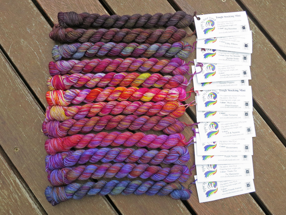 Twelve variegated mini skeins of yarn in shades of grey, mauve and purple to blue with white labels are arranged from darkest grey at top to lightest grey then through mauve to purple then navy blue at bottom, lying on a brown wooden background.