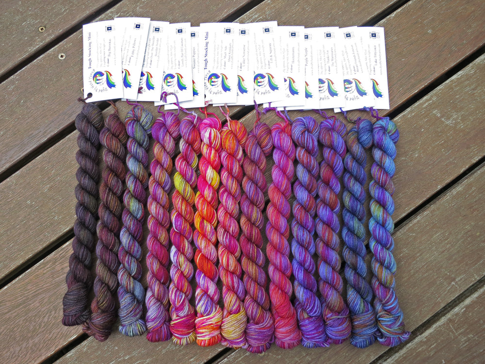 Thirteen variegated mini skeins of yarn in shades of grey, mauve and purple to blue with white labels are arranged from darkest grey at left to lightest grey then through mauve to purple then blues at right, lying on a brown wooden background.